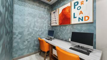 A modern office space with two desks, each with a computer and keyboard, and two orange chairs. The walls have a geometric pattern, and there are framed artworks above the desks.