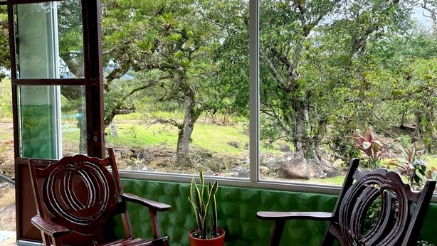 A room with a large window, two rocking chairs, a small table with a potted plant, and a chair. - Miravalles Volcano House (Mogote de Bagaces)
