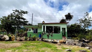 A single-story building with a green exterior and a red roof, surrounded by a grassy area with rocks and trees. - Miravalles Volcano House (Mogote de Bagaces)