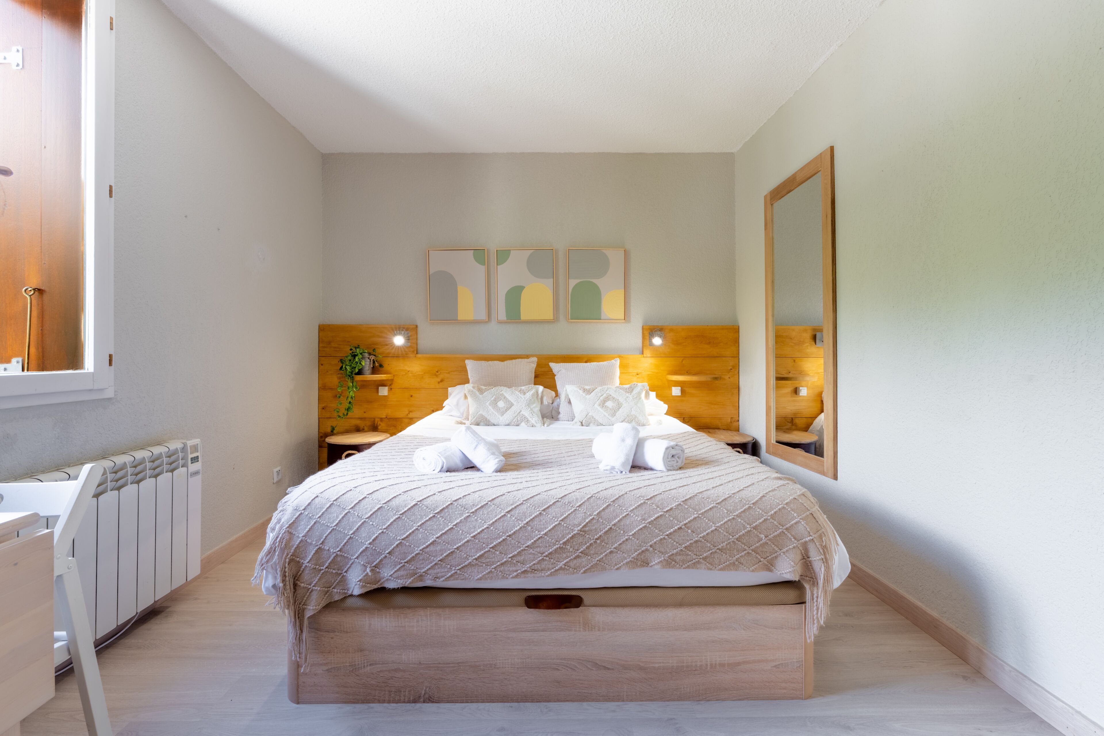 A bedroom with a bed, bedside tables, a radiator, a mirror, and abstract wall art.
