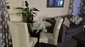 Een woonkamer met een ronde glazen tafel, twee witte stoelen, een donkergrijze bank met gekleurde kussens, een kamerplant en een raam met gordijnen.