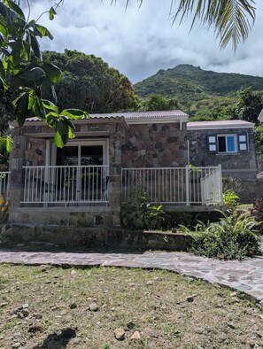 Un bâtiment en pierre doté d’un balcon, entouré de verdure et adossé à un paysage montagneux.