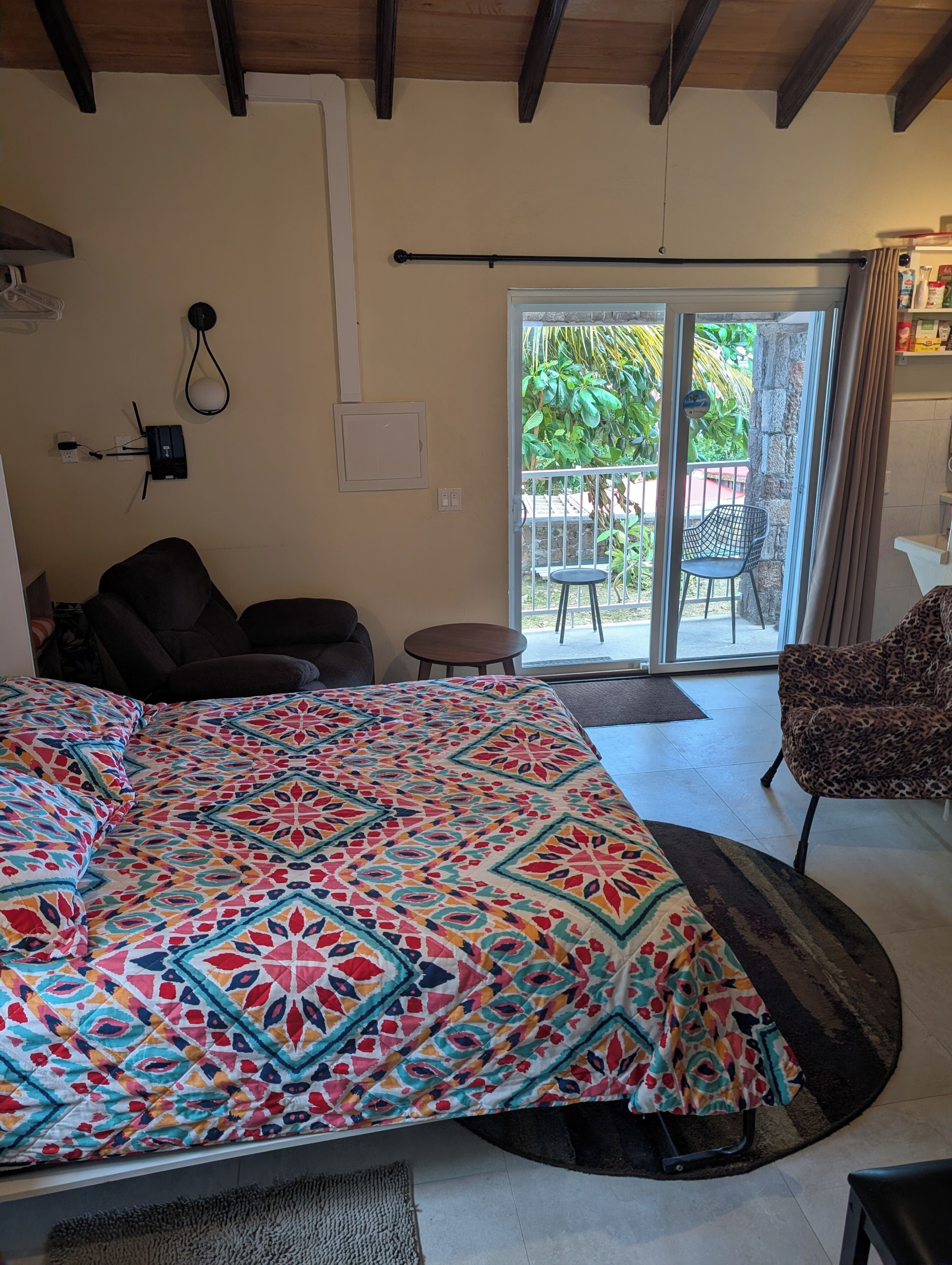 Une pièce comprenant un grand lit, un canapé, un fauteuil, une petite table et une porte-fenêtre coulissante donnant sur un espace extérieur agrémenté de plantes et d’un fauteuil.