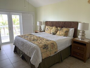 A bedroom with a large bed, two bedside tables with lamps, a sliding glass door leading to a balcony, and a view of palm trees.