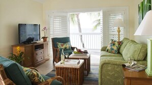 Una sala de estar con televisor, una mesa de centro, un sofá y una silla. Hay una ventana con persianas y vistas a palmeras afuera.