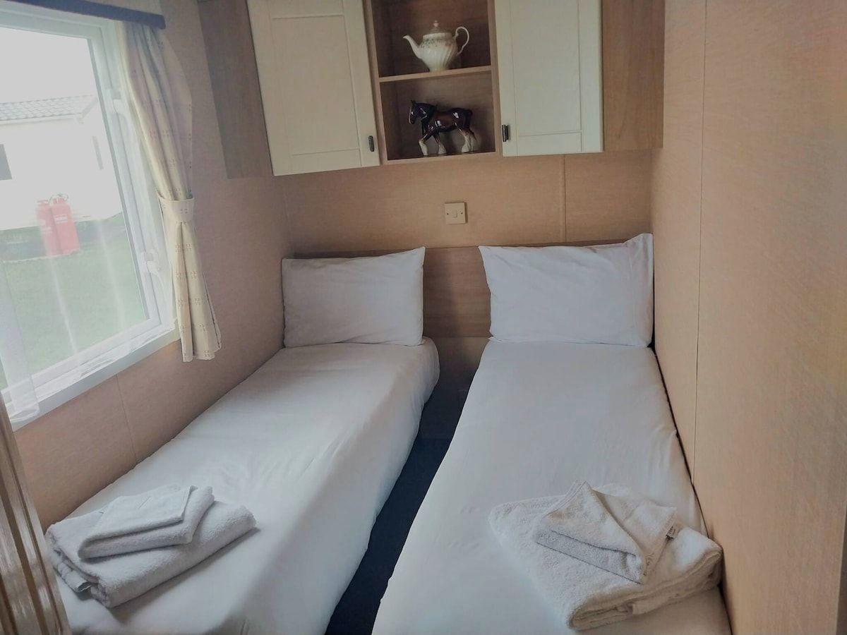 A room with two beds, a window with curtains, and a shelf with a teapot and horse figurines.