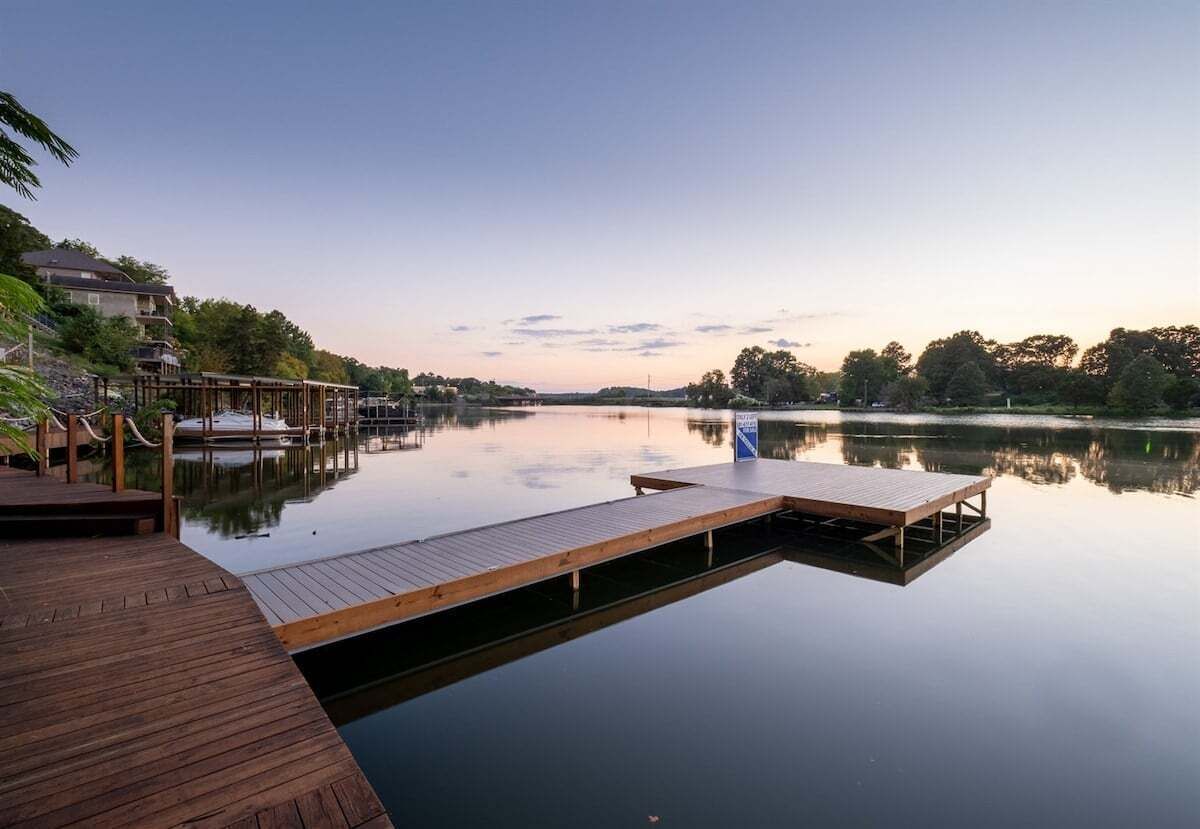 Een rustig meer met een houten steiger die in het water reikt, omgeven door bomen en een huis aan de linkerkant.