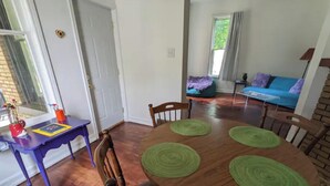 Ein Essbereich mit einem Holztisch, vier Stühlen und grünen Platzsets. Im Vordergrund steht ein blauer Tisch mit einer roten Vase und einem gelben Platzset.