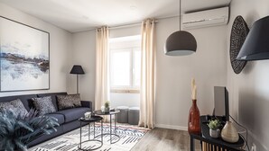 Een moderne woonkamer met een donkere bank, een kamerkleed met dessin, een bijzettafeltje met een lamp en een groot raam met gordijnen.