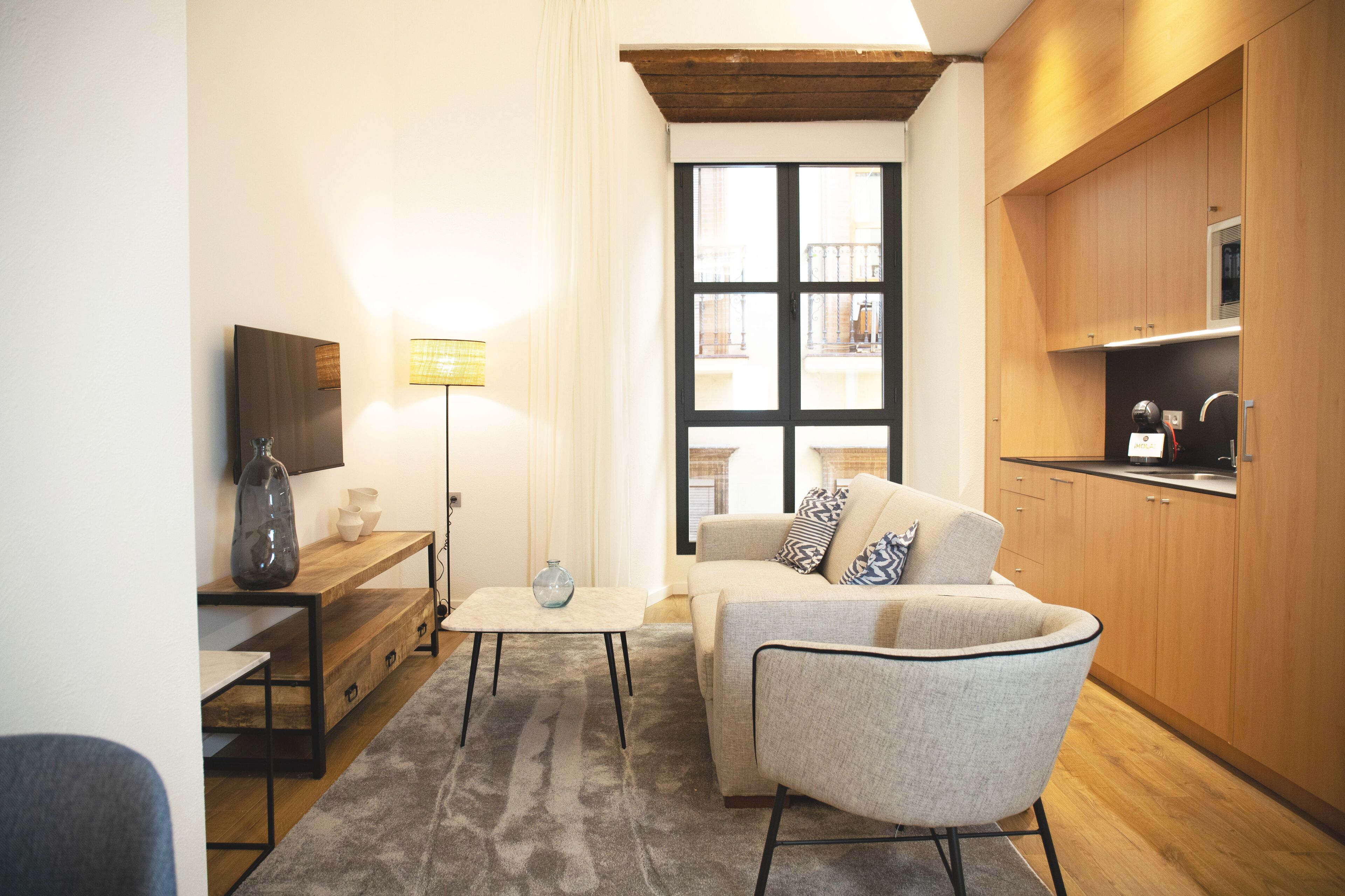 A modern living room with a sofa, coffee table, and lamp. There is a kitchen area with wooden cabinets and a window with blinds.