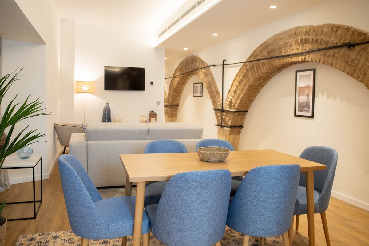 A modern living room with a wooden dining table, blue upholstered chairs, a grey sofa, and a TV mounted on the wall.