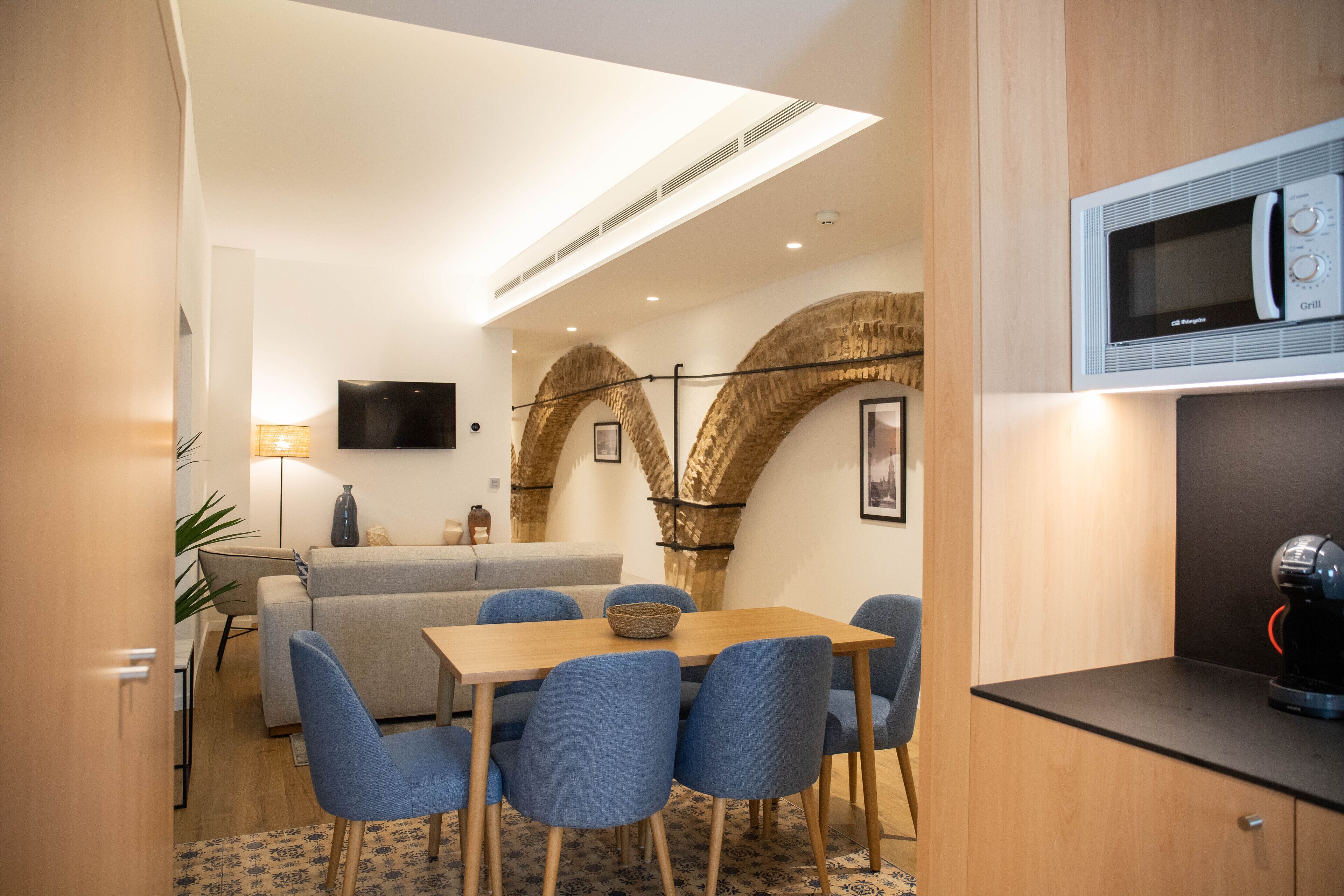 A modern living room with a sofa, dining table, and chairs. There is a flat-screen TV mounted on the wall, a microwave on the counter, and a coffee machine.