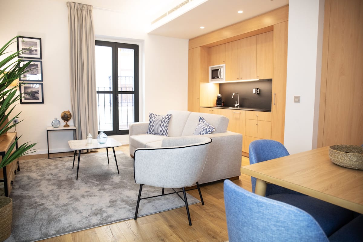 A modern living room with a sofa, a coffee table, and a dining table. There is a kitchen area with wooden cabinets and a microwave.