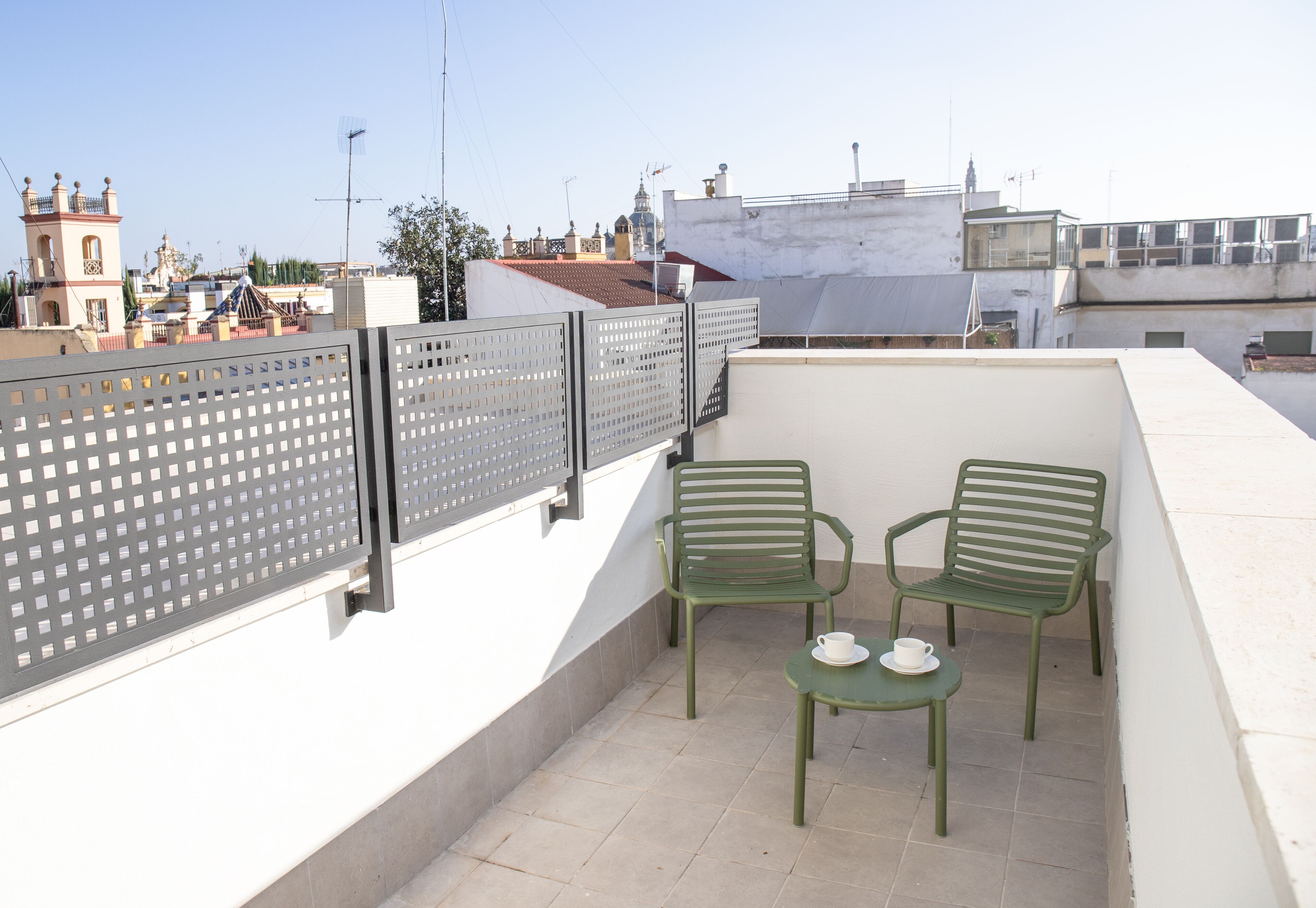 A rooftop terrace with two green chairs and a small table, a white railing, and a view of the cityscape.