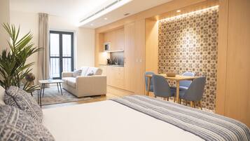A modern hotel room with a large bed, a sofa, a dining area, and a kitchenette.