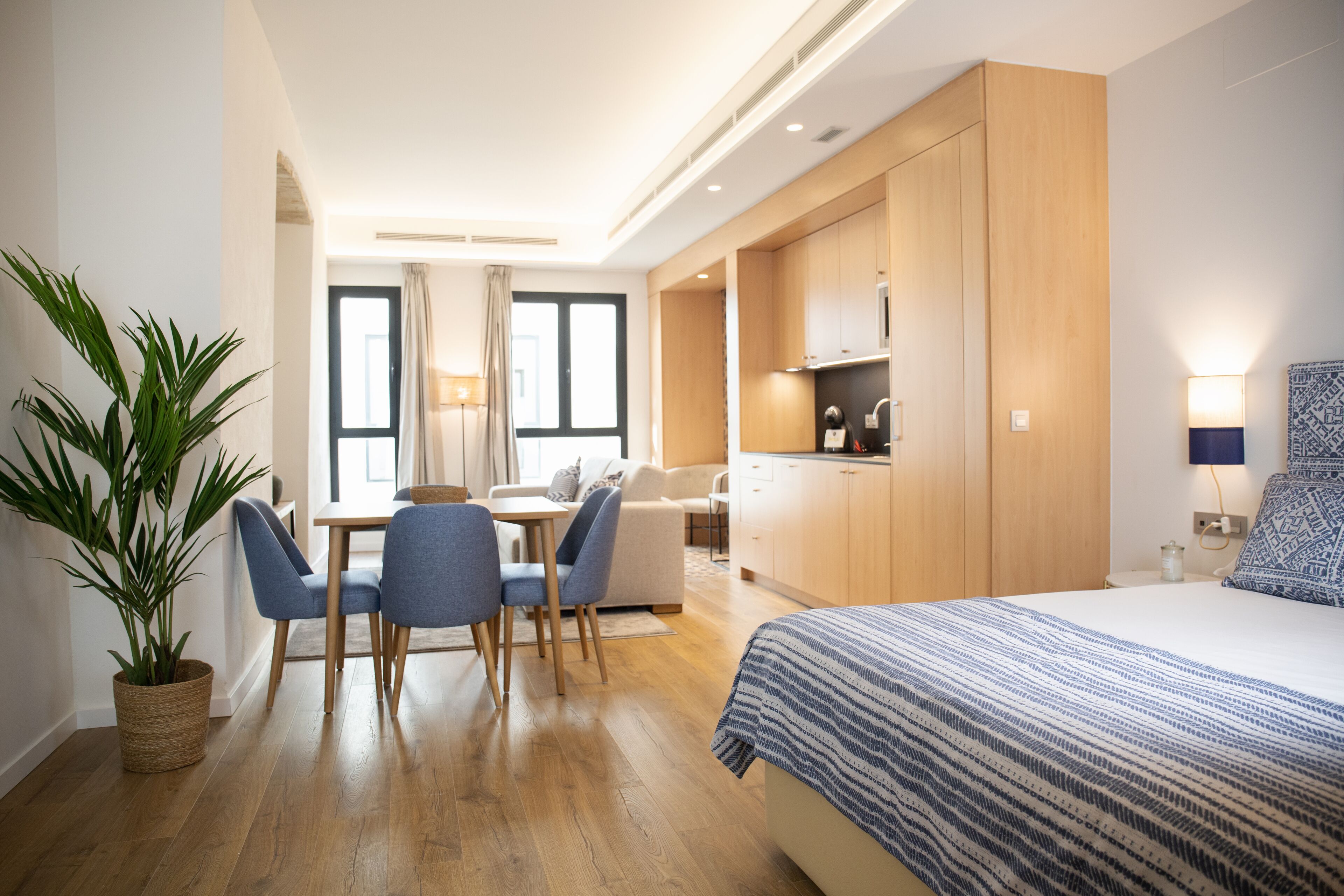 A modern hotel room with a bed, a dining area with chairs and a table, a kitchenette, and a large potted plant.