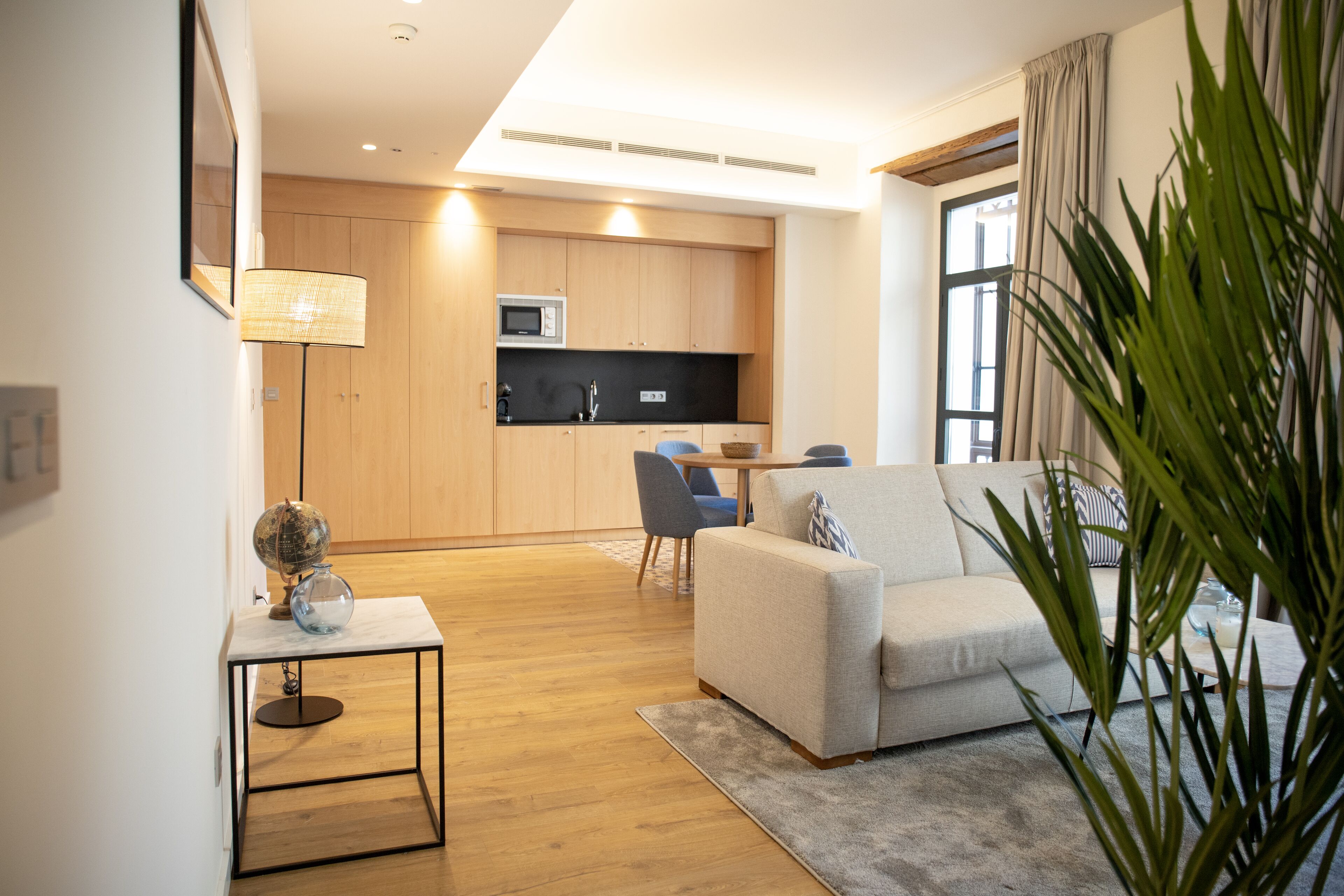A modern living room with a sofa, a coffee table, and a lamp. There is a kitchen area with wooden cabinets and a microwave.