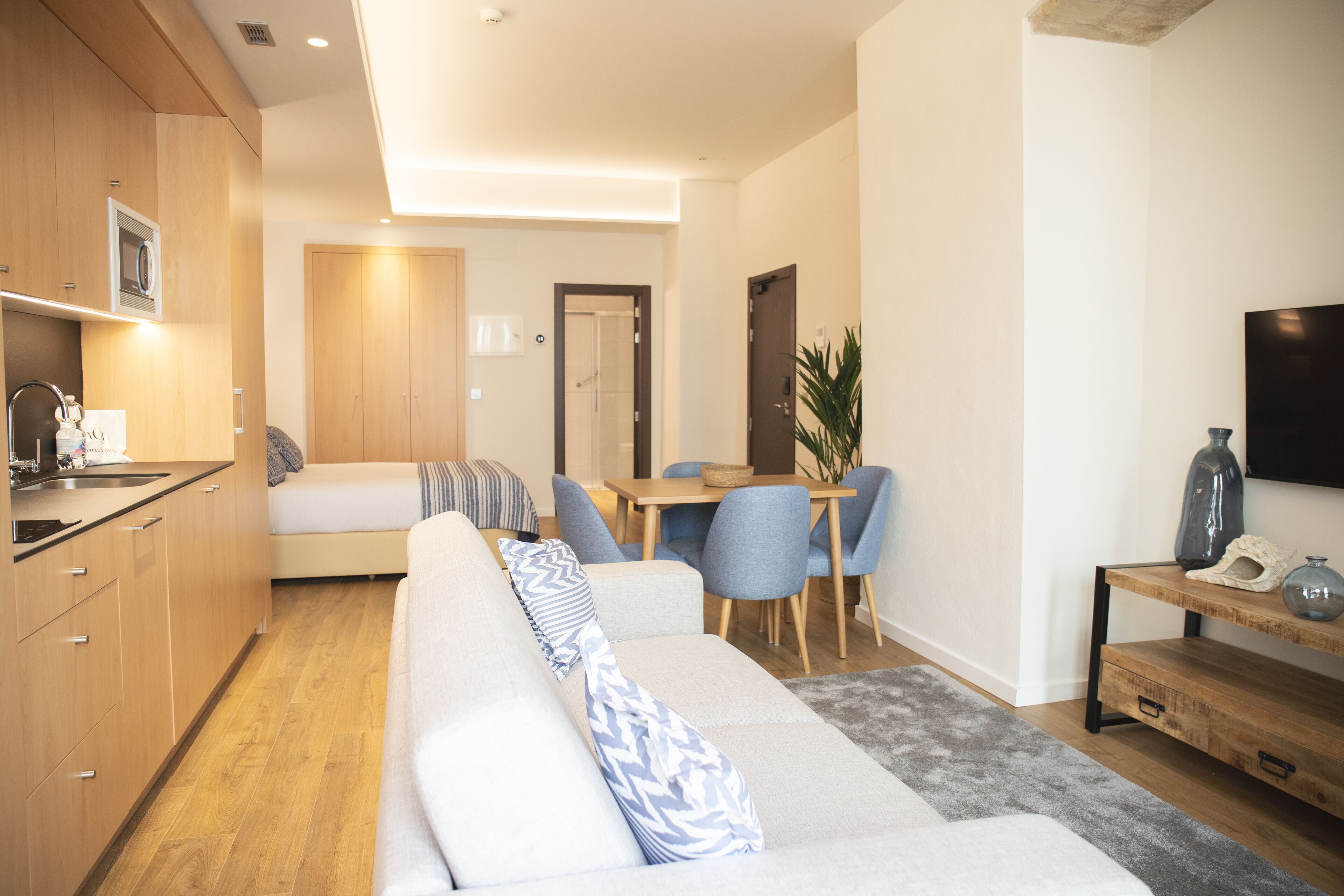 Una sala de estar moderna con un sofá, un comedor con sillas, una cocina con armarios de madera y un televisor montado en la pared.