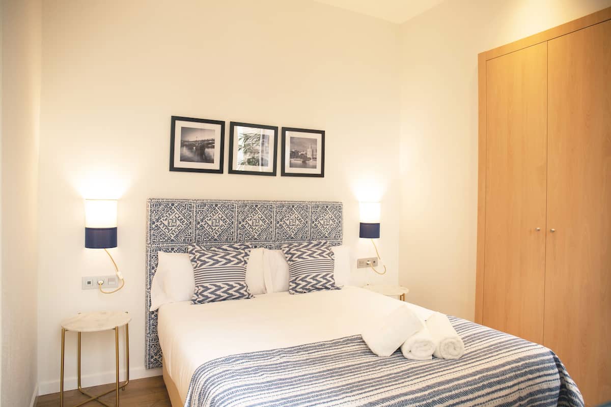 A hotel room with a bed, two wall-mounted lamps, a patterned headboard, and a wooden wardrobe.