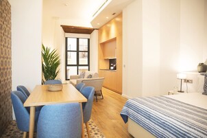 A modern hotel room with a wooden dining table, blue chairs, a sofa, a bed with a striped blanket, a potted plant, and a kitchenette in the background.