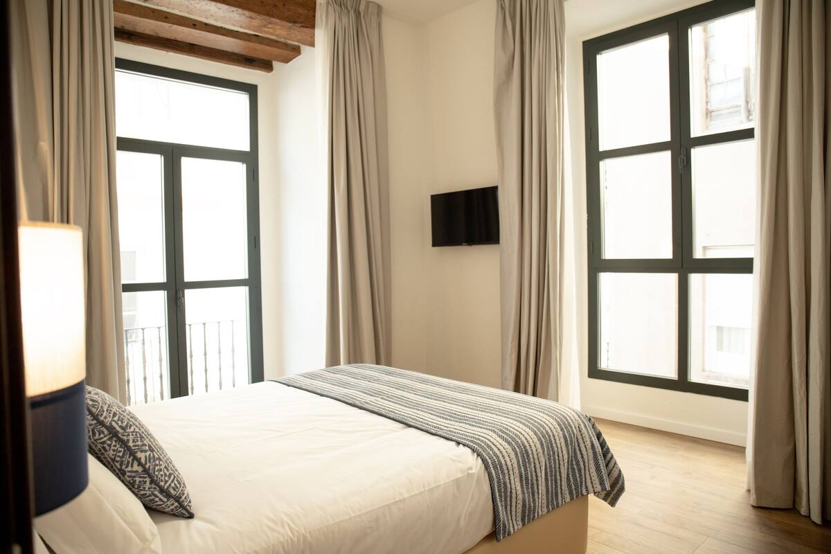 A bedroom with a bed, curtains, a TV, and large windows.