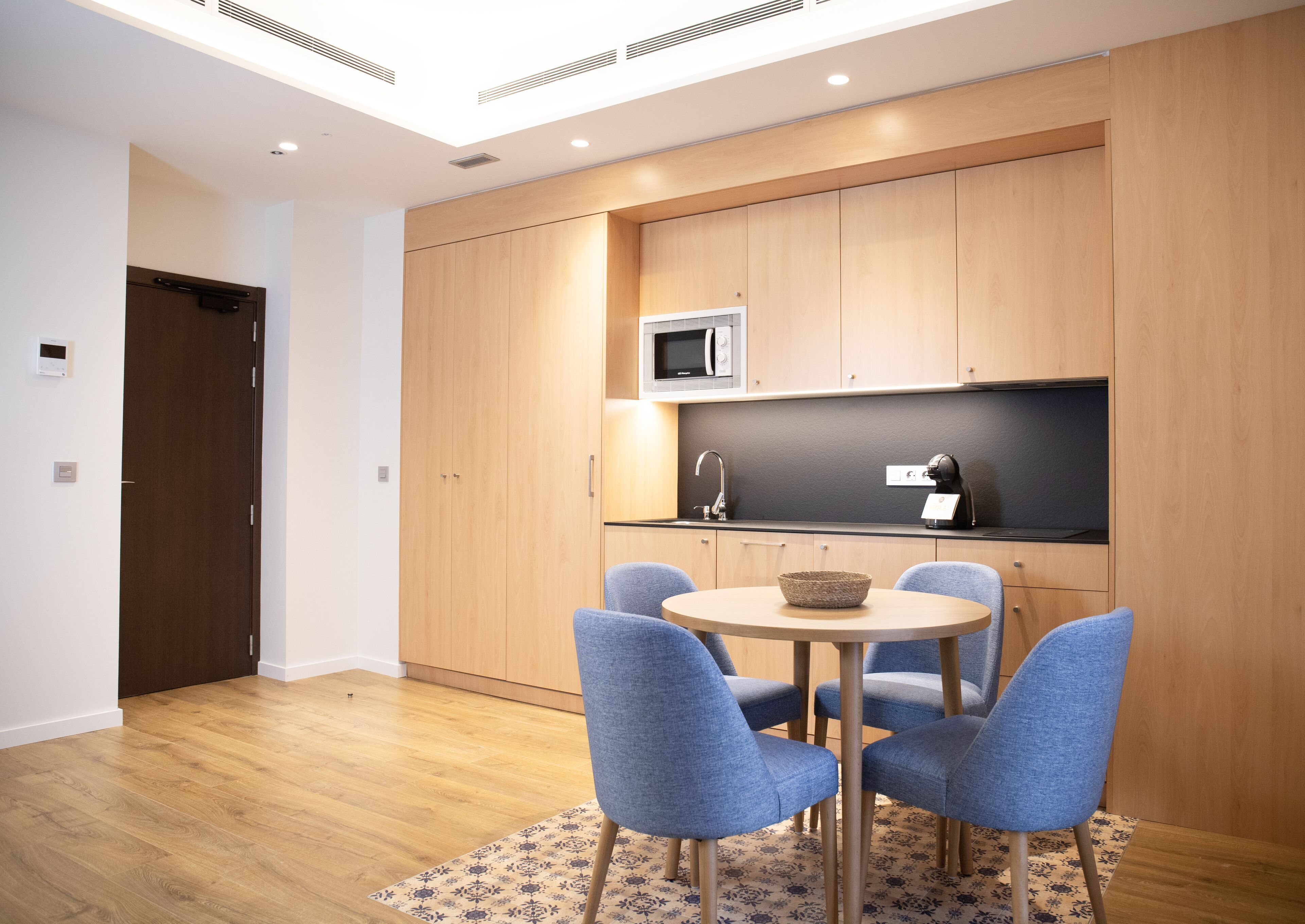 A modern kitchen with wooden cabinets, a microwave, and a sink. There is a round table with four blue chairs and a woven basket on it.