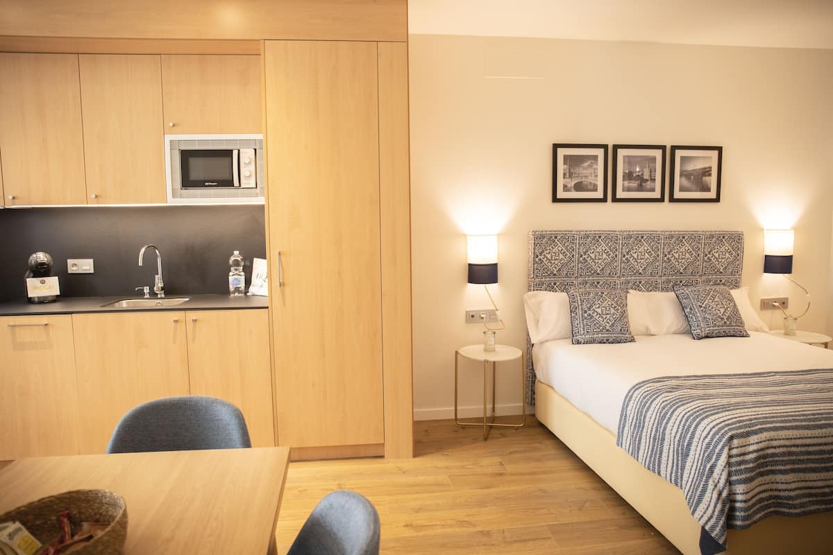 A compact hotel room with a kitchenette, a bed with patterned pillows, bedside lamps, and a small side table.