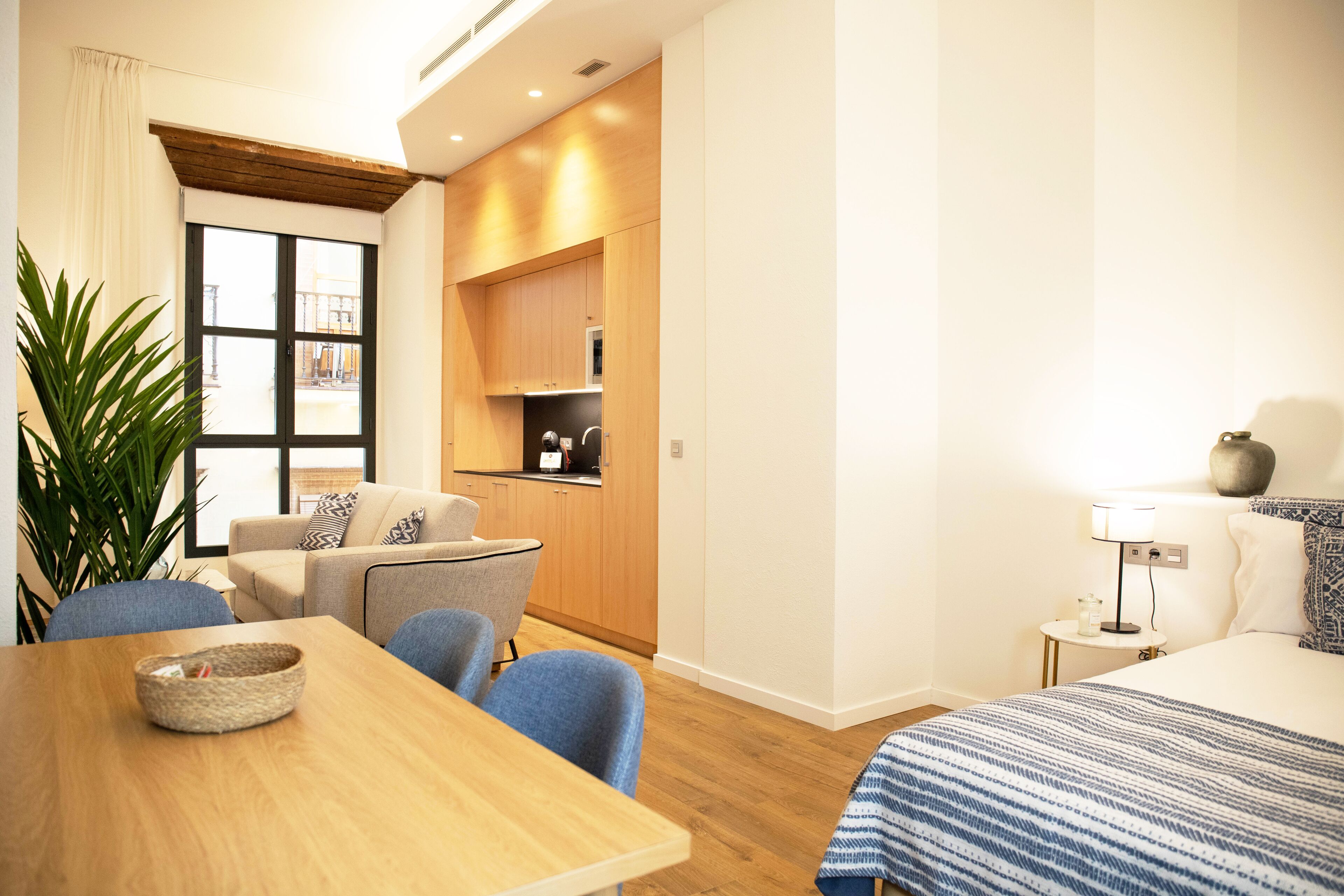 A modern hotel room with a wooden dining table, a sofa, a small kitchen area, and a bed with a blue and white striped bedspread.