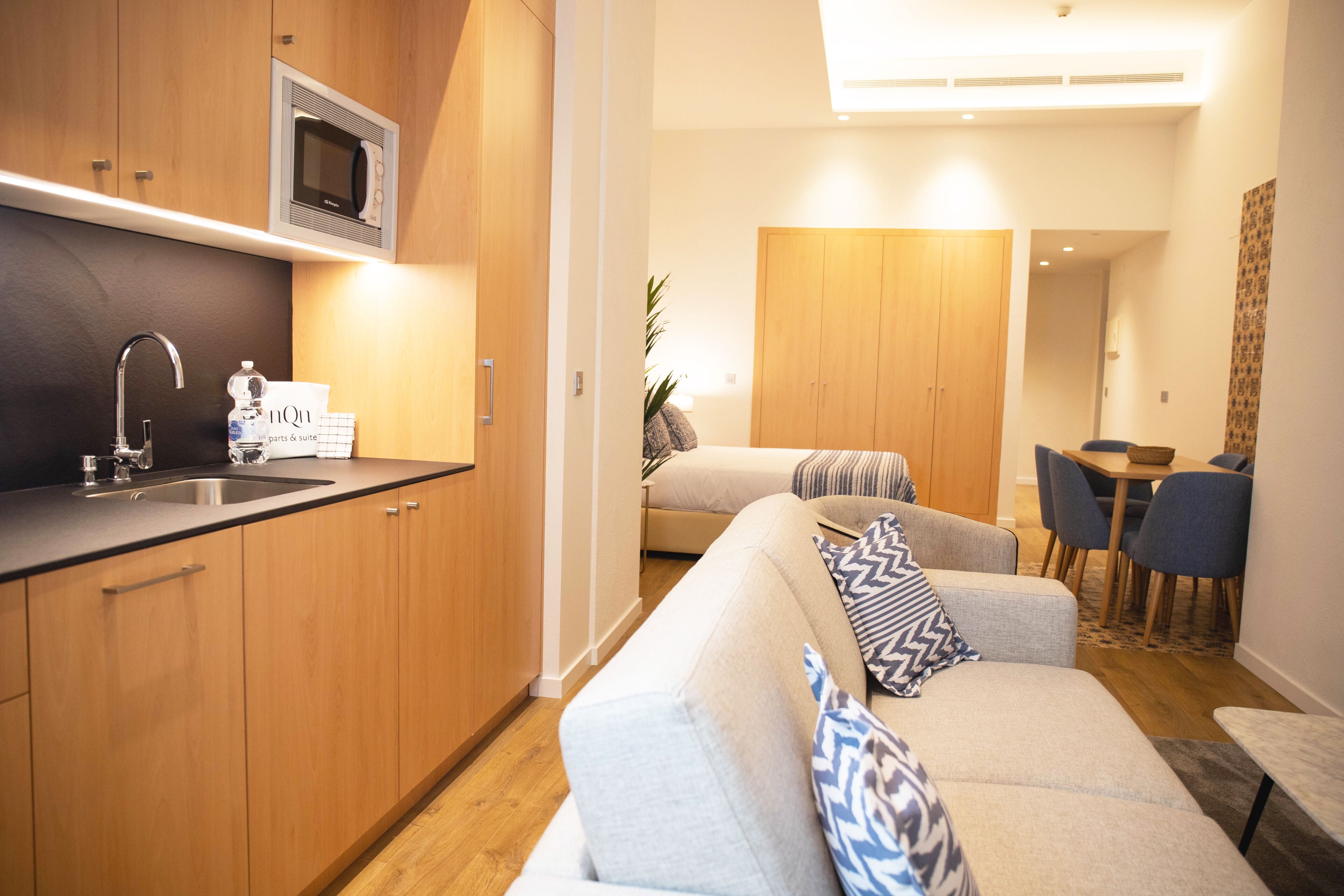 Un espace de vie compact comprenant un coin cuisine, un canapé avec des coussins à motifs, un coin repas avec des chaises et une armoire en bois.