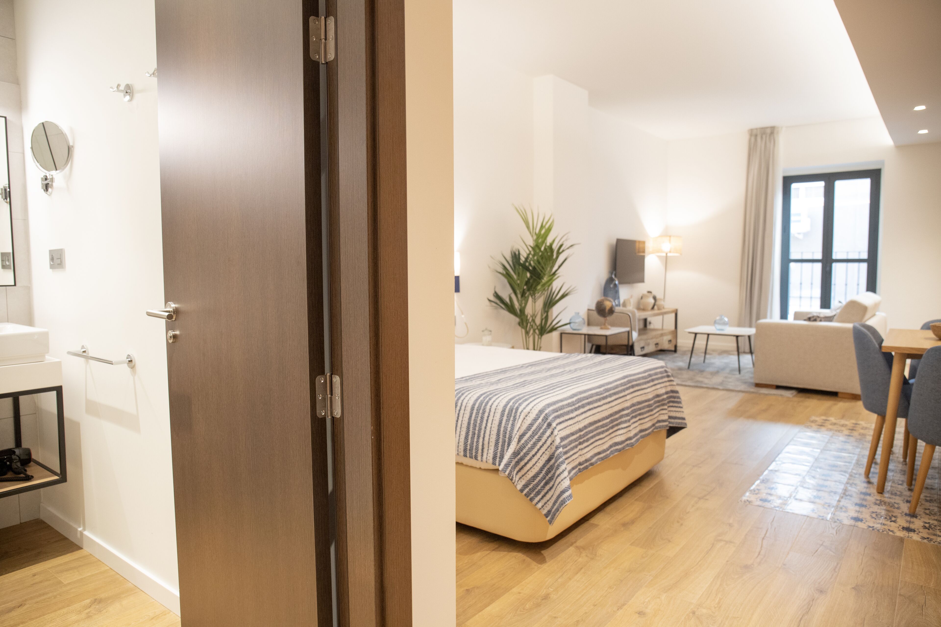 A modern hotel room with a wooden door, a bed with a striped blanket, a small table with a lamp, a chair, and a large window.