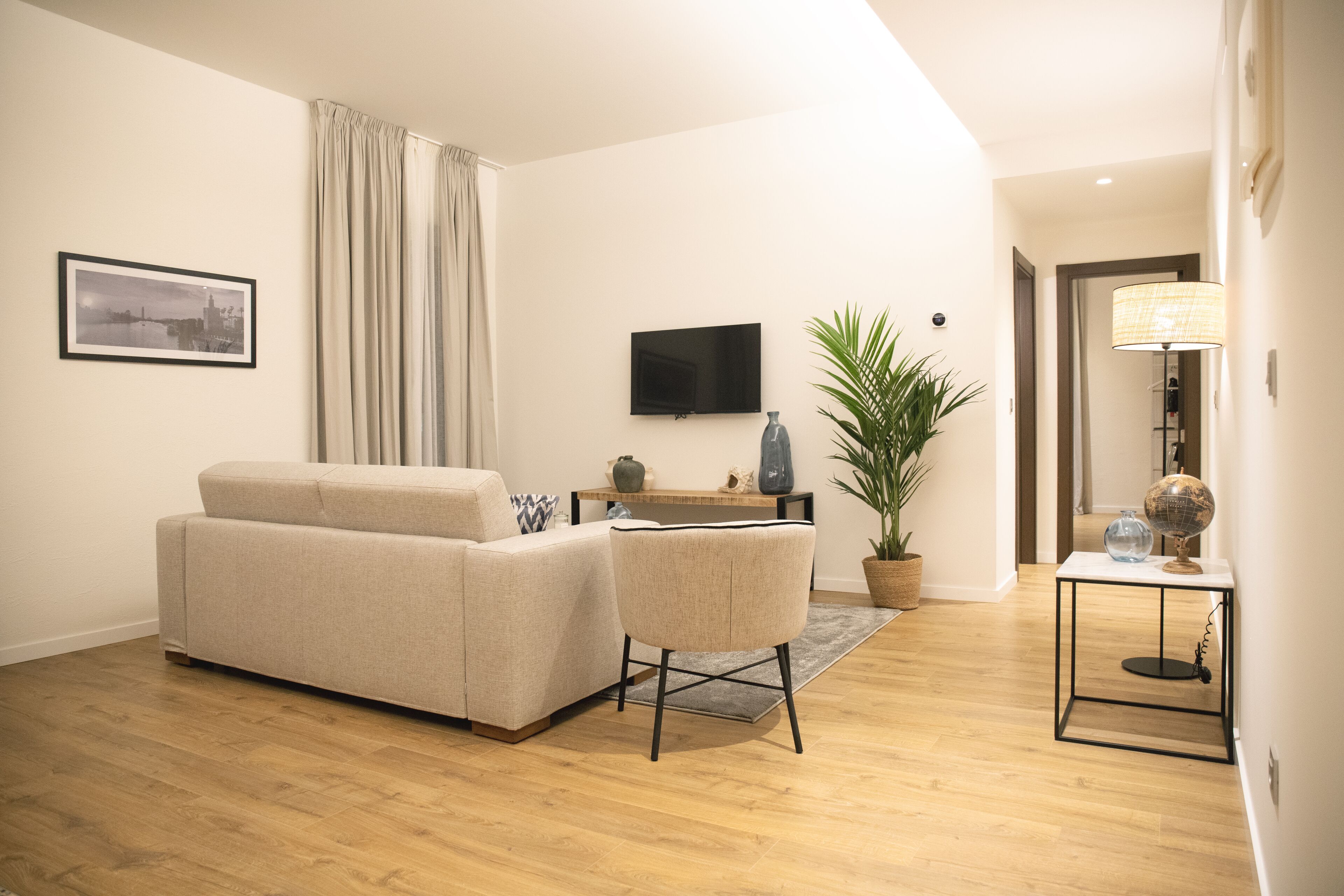Una sala de estar moderna con un sofá, una silla, un televisor, una planta y una lámpara sobre una mesa.