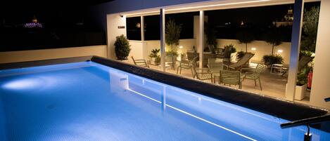 A swimming pool with a clear blue water surface, surrounded by a covered patio area with chairs and potted plants.