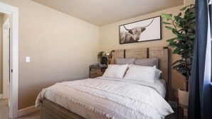 A bedroom with a wooden bed, a nightstand, a wall-mounted lamp, a large plant, and a framed picture. - Patio w/ fire pit home ✶ 10min to DT → Has it all! (Portland)