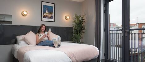 Quarto de hotel moderno com uma cama grande, vista da cidade através de uma varanda, e um quadro na parede.