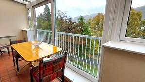 A balcony with a wooden table and chairs, a view of trees and mountains, and a railing. - Loch Rannoch Highland Club Apartment 16 (Kinloch Rannoch)