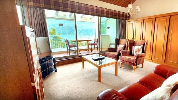 A living room with a red sofa, a wooden coffee table, and a plaid armchair. There is a view of a balcony through large windows. - Loch Rannoch Highland Club Apartment 16 (Kinloch Rannoch)