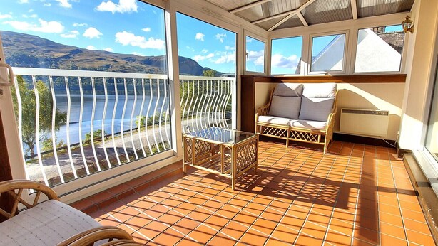 A sunlit balcony with a view of a lake and mountains, furnished with wicker chairs and a small table. - Loch Rannoch Highland Club Studio 2 (Kinloch Rannoch)