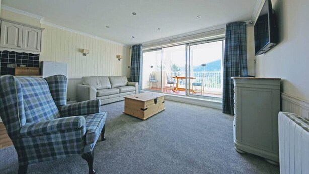 A spacious living room with a sofa, armchair, and a television mounted on the wall. - Loch Rannoch Highland Club Apartment 9 (Kinloch Rannoch)