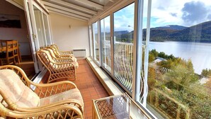 Un balcón con sillas de mimbre y una mesa, con vista a un lago y colinas.