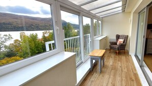 Ein Balkon mit Holztisch und kariertem Stuhl, mit Blick auf einen See und Hügel.