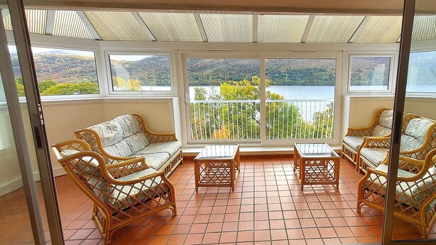 Un balcon ensoleillé, agrémenté de meubles en osier, doté d’un sol carrelé et offrant une vue panoramique sur les collines et l’eau.