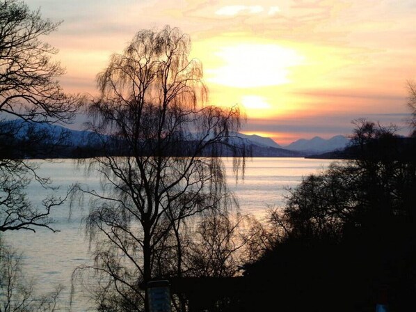 A serene lakeside view with a sunset, trees, and distant mountains. - Loch Rannoch Highland Club Studio 1 (Kinloch Rannoch)