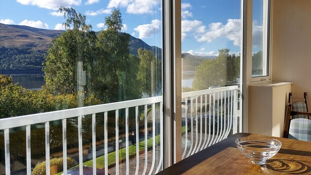 A balcony with a clear view of a lake, mountains, and a parking area. - Loch Rannoch Highland Club Apartment 15 (Kinloch Rannoch)