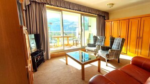 A living room with a glass-top coffee table, a brown leather sofa, and plaid armchairs. There is a wooden cabinet and a refrigerator in the background. - Loch Rannoch Highland Club Apartment 15 (Kinloch Rannoch)