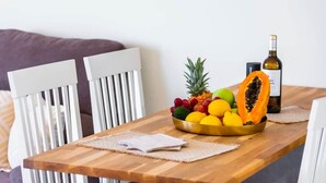 Una mesa de madera para comer con un frutero que contiene piña, mango y otras frutas, junto con una botella de vino.