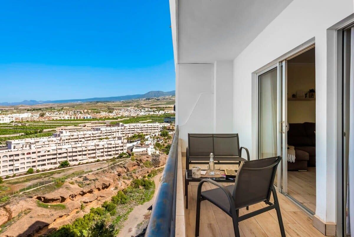 Un balcon avec une table et des chaises, donnant sur un quartier résidentiel avec des immeubles et un ciel dégagé.