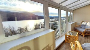 Un balcon agrémenté de meubles en osier, d’une table en verre et offrant une vue sur un lac et des montagnes.