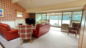 A living room with a red sofa, a plaid armchair, and a television. There is a large window with a view of the outdoors. - Loch Rannoch Highland Club, Highland  Lodge 45 (Kinloch Rannoch)