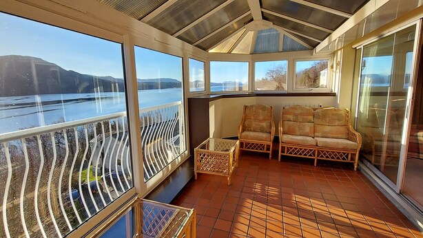 A sunroom with large windows overlooking a body of water, wicker furniture, and a tiled floor. - Loch Rannoch Highland Club, Highland  Lodge 45 (Kinloch Rannoch)