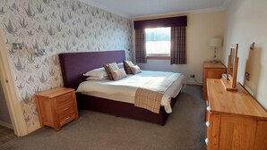 A bedroom with a bed, wooden nightstands, a dresser, a window with curtains, and a wall with a floral pattern. - Loch Rannoch Highland Club, Highland Lodge 35 (Kinloch Rannoch)
